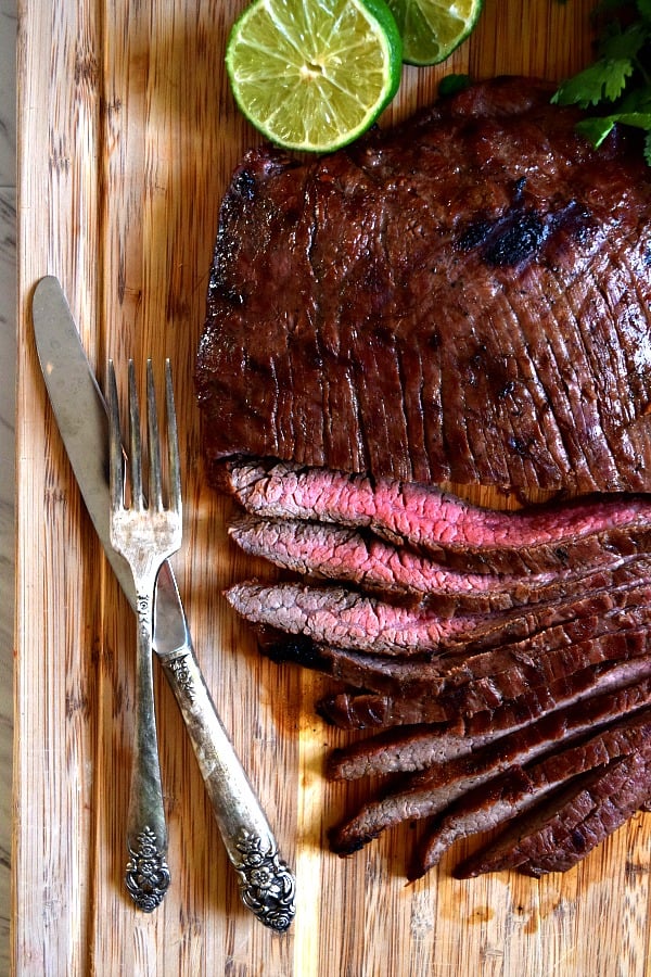 Grilled Flank Steak on cutting board half cut into strips with a fork, knife, & limes