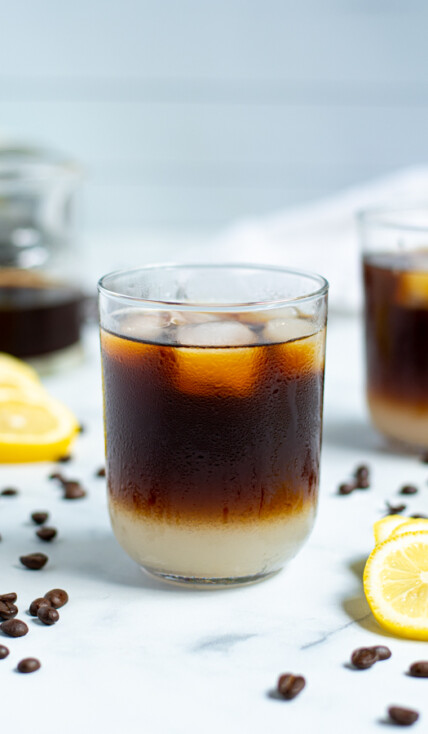 drinking glass with coffee beans and lemon slices surrounding it filled with layered lemonade on the bottom and iced coffee on top with ice