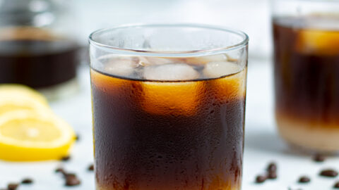 drinking glass with coffee beans and lemon slices surrounding it filled with layered lemonade on the bottom and iced coffee on top with ice