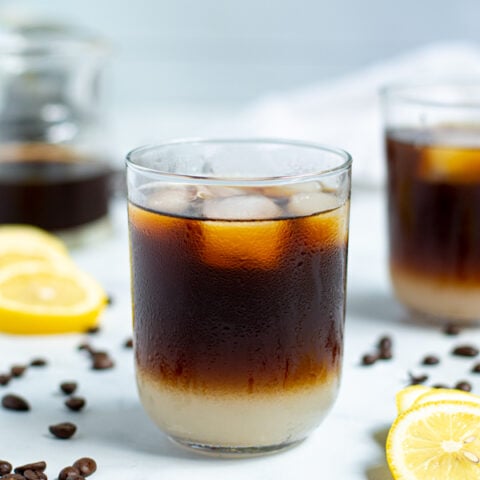 drinking glass with coffee beans and lemon slices surrounding it filled with layered lemonade on the bottom and iced coffee on top with ice