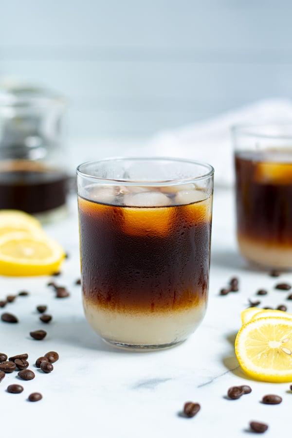 drinking glass with coffee beans and lemon slices surrounding it filled with layered lemonade on the bottom and iced coffee on top with ice