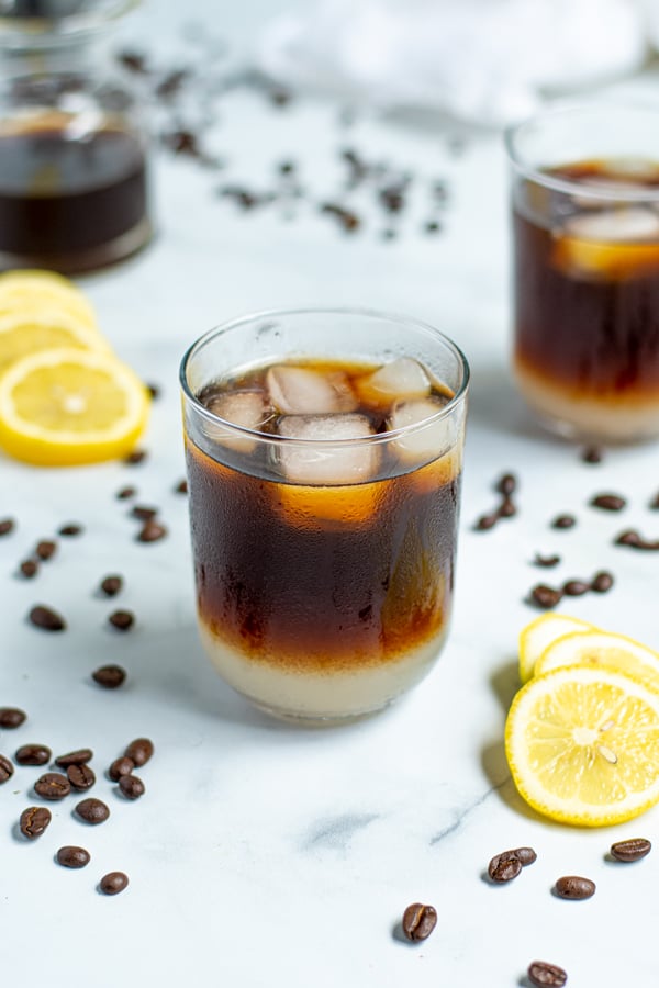 in the foreground is a drinking glass layered with lemonade on the bottom and cold brew coffee on top with ice