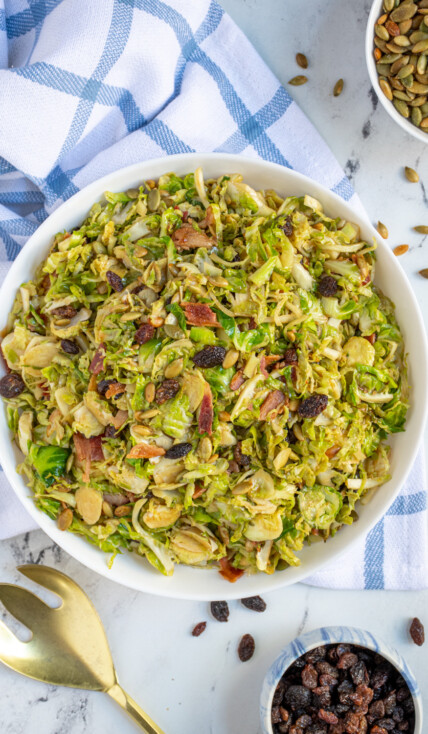 Overhead view of a large white bowl full of warm balsamic brussel sprout salad with pumpkin seeds, raisins, and bacon.
