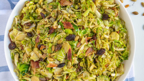 Overhead view of a large white bowl full of warm balsamic brussel sprout salad with pumpkin seeds, raisins, and bacon.