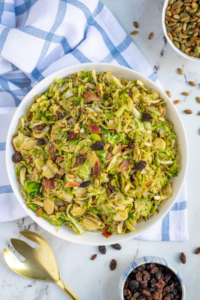 Overhead view of a large white bowl full of warm balsamic brussel sprout salad with pumpkin seeds, raisins, and bacon.