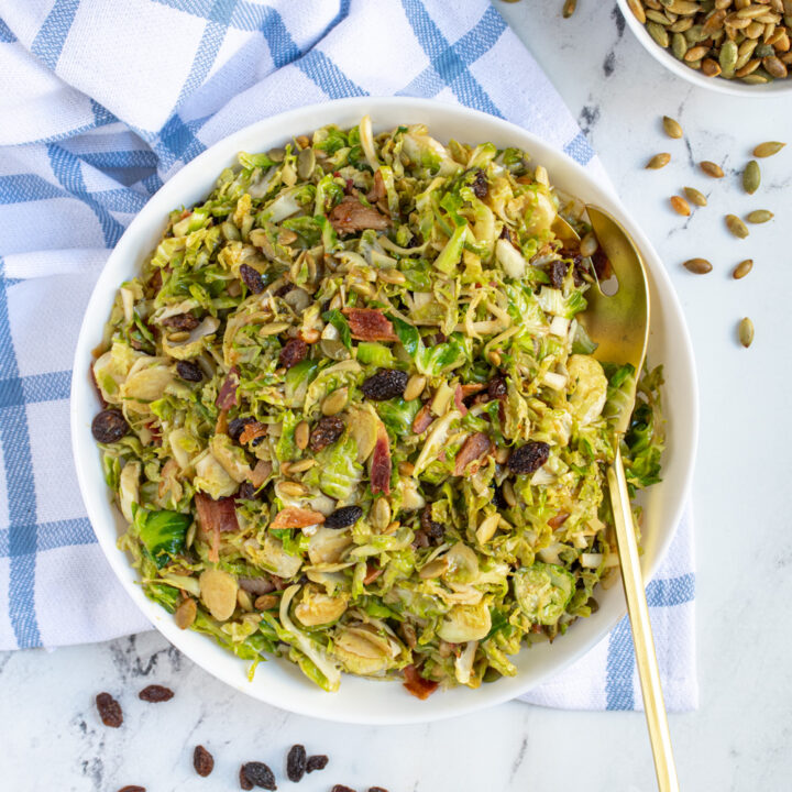 White serving bowl and gold serving spoon with a warm brussel sprout bacon salad garnished with raisins and pumpkin seeds.