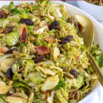 Pinterest Pin with text 'Warm Balsamic Brussel Sprout Salad with Bacon', image of a serving bowl full of shaved brussel sprout salad with bacon, raisins, and pumpkin seeds.