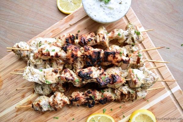 Grilled Greek Chicken Kabobs stacked up on a wood cutting board next to slices lemons