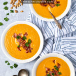 Pinterest Pin with text overlay, Roasted Butternut Squash Soup, image of three bowl on a table top filled with soup and topped with bacon, sitting next to a kitchen towel and spoons.