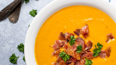 Overhead view of a white bowl full of roasted butternut squash and carrot soup garnished with bacon crumbles and fresh parsley.