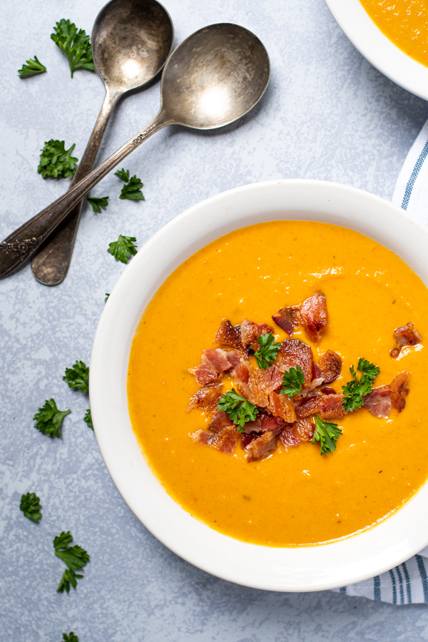 Overhead view of a white bowl full of roasted butternut squash and carrot soup garnished with bacon crumbles and fresh parsley.