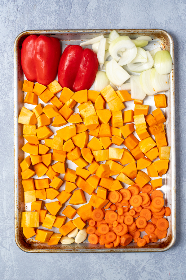 Overhead view of a sheet pan with freshly diced butternut squash, carrots, and onion with halved red bell peppers.
