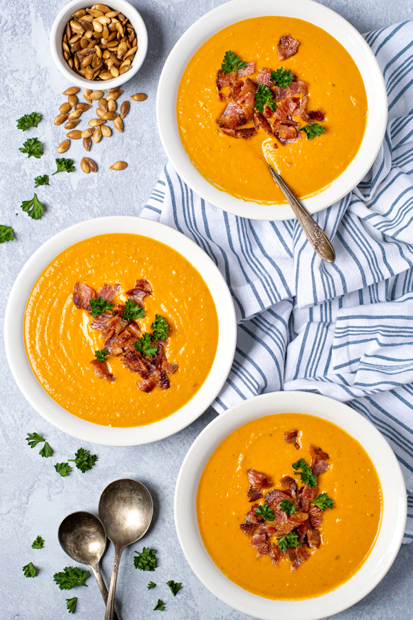 Flatly of three white bowls filled with roasted butternut squash soup garnished with bacon sitting next to a kitchen towel, spoons, and baked squash seeds.