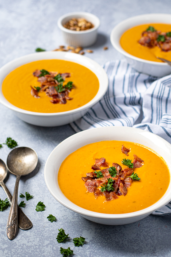 Three bowls placed on a table top next to spoons, in the bowl is curried butternut squash soup garnished with bacon crumbles.
