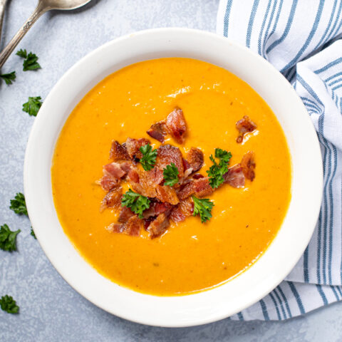 Close up on a bowl filled with butternut squash soup on a table top next to spoons and a blue kitchen towel.