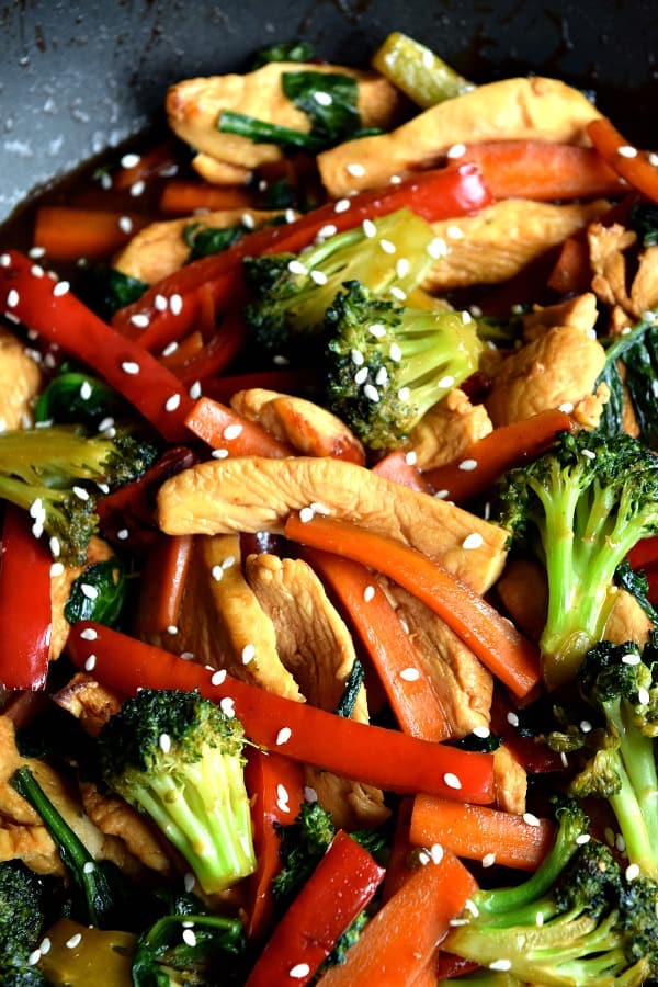 a close up of chicken stir fry with carrots, peppers, and broccoli, topped with sesame seeds