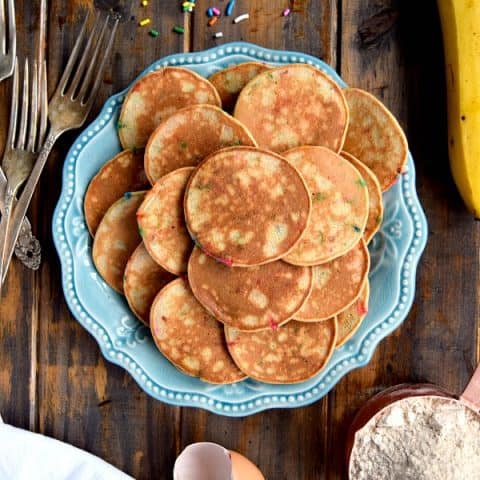Mini Whole Wheat Banana Pancakes on a blue plate surrounded by sprinkles, a cup of flour, and a banana