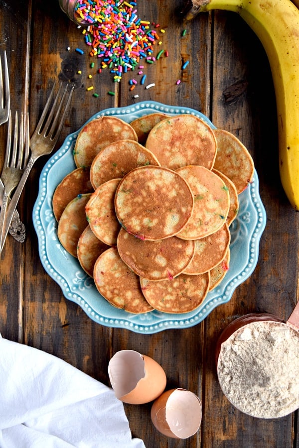 Mini Whole Wheat Banana Pancakes on a blue plate surrounded by sprinkles, a cup of flour, and a banana