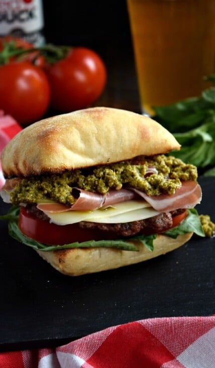 a close up of a prosciutto pesto burger on a black plate with a beer