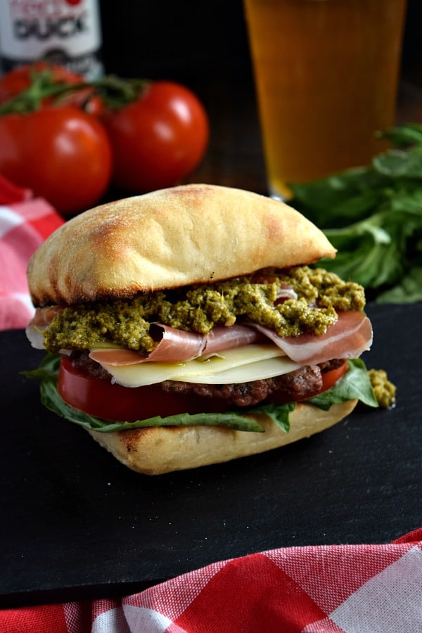 a close up of a prosciutto pesto burger on a black plate with a beer
