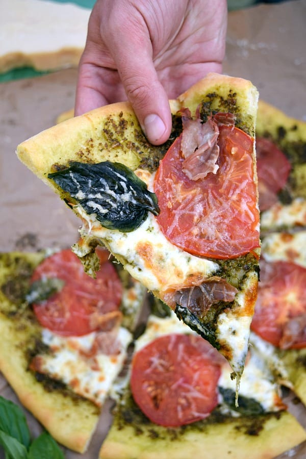 Pesto Pizza with Prosciutto, Tomatoes, and Mozzarella slice being held by a hand ready to be eaten