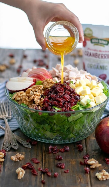 Apple Cider Vinaigrette being poured onto an Apple Cranberry Salad with turkey, walnuts, and cheddar cheese