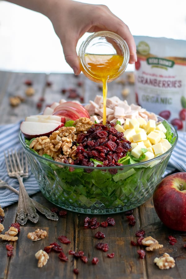 Apple Cider Vinaigrette being poured onto an Apple Cranberry Salad with turkey, walnuts, and cheddar cheese