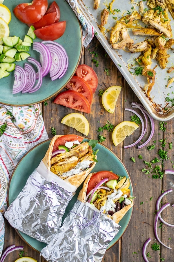 pair of wrapped slow cooker chicken shawarmas on a plate next to a sheet tray of shredded chicken 