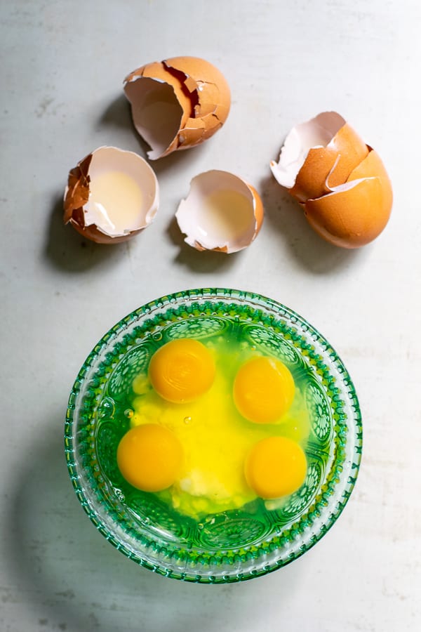 4 cracked eggs in a blue bowl with the brown shells sitting next to it 