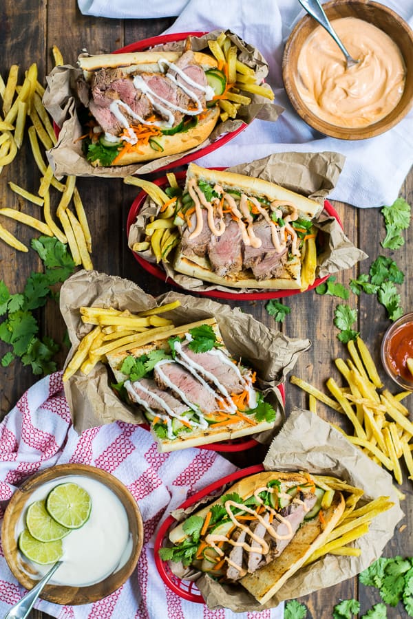 four serving baskets filled with fries and grilled pork banh mi's on a table top with flavorful mayo sitting in bowls