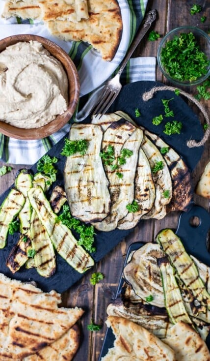 a spread of grilled zucchini, eggplant, naan, and hummus over a wooden background