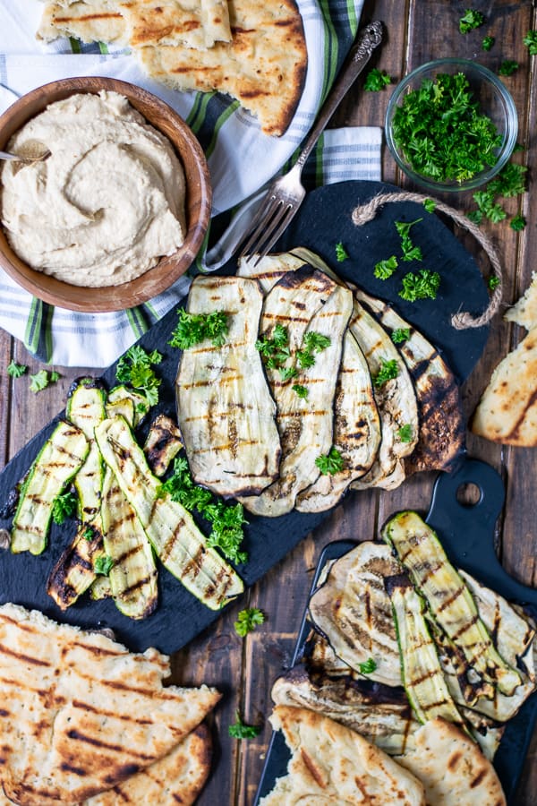 a spread of grilled zucchini, eggplant, naan, and hummus over a wooden background
