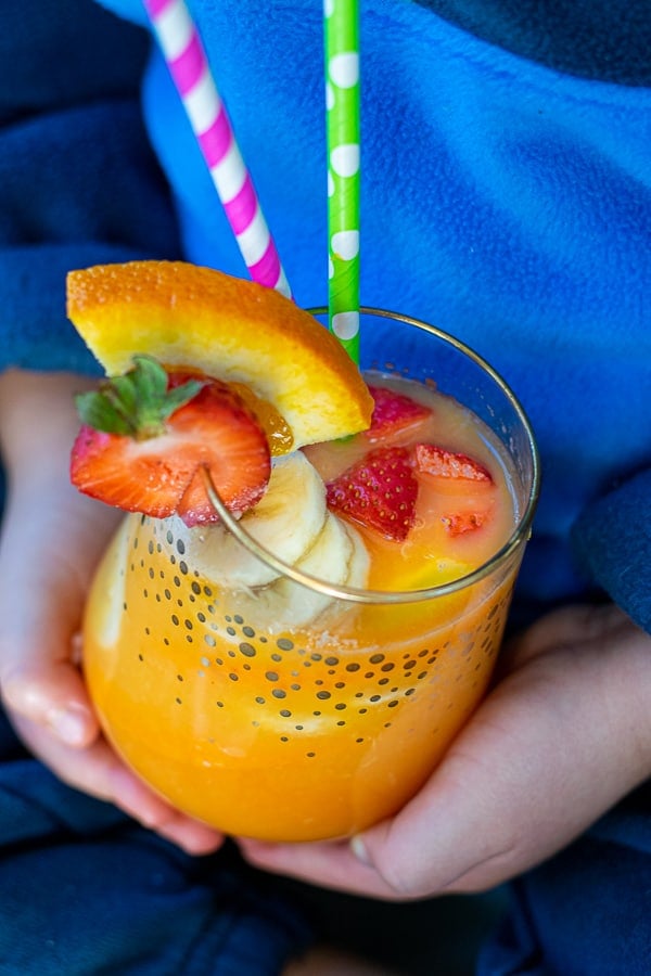 kids hands holding a glass of orange juice filled with colorful fruits