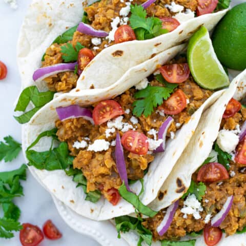 three red lentil tacos with vegetarian taco meat on a white plate with garnishes