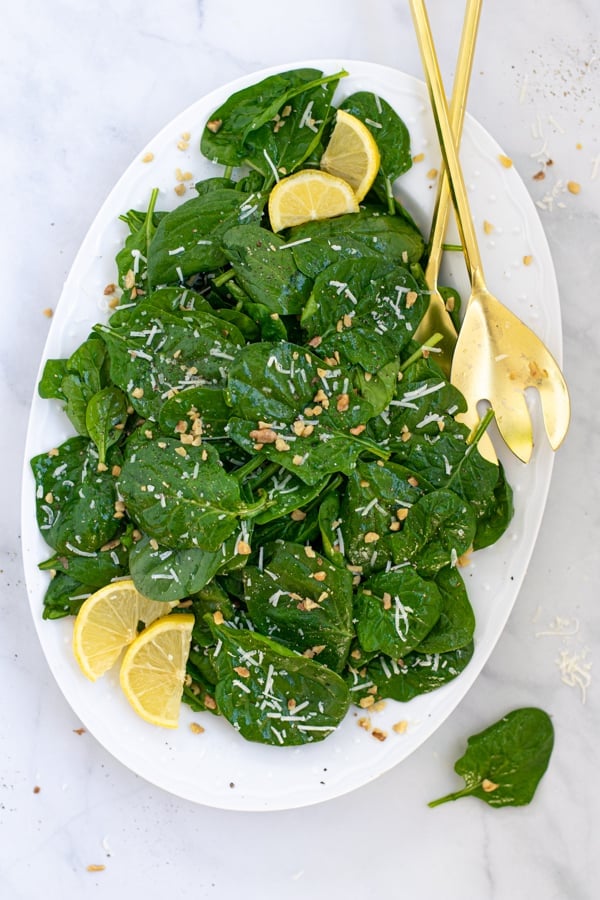 overhead view of a simple spinach salad on a white platter with gold serving utensils