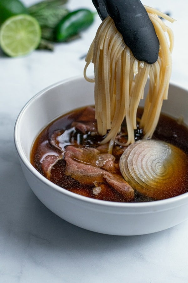 noodles being placed into the soup bowl with a pair of tongs