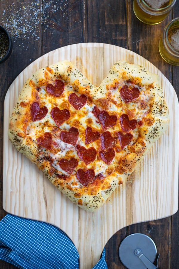heart shaped pizza just pulled out of the oven on a pizza paddle with a pizza cutter and a couple of beers