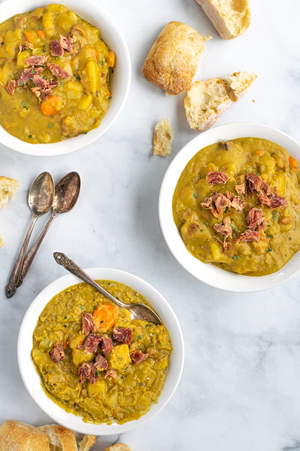 3 bowls of split pea soup on a table with spoons and bread rolls
