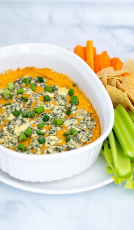 easy Buffalo Chickpea Dip in a small white baking dish with blue cheese and green onions with chips and vegetables for serving
