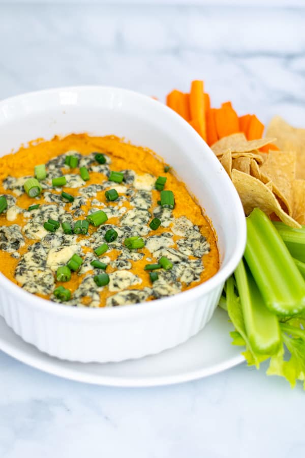 easy Buffalo Chickpea Dip in a small white baking dish with blue cheese and green onions with chips and vegetables for serving