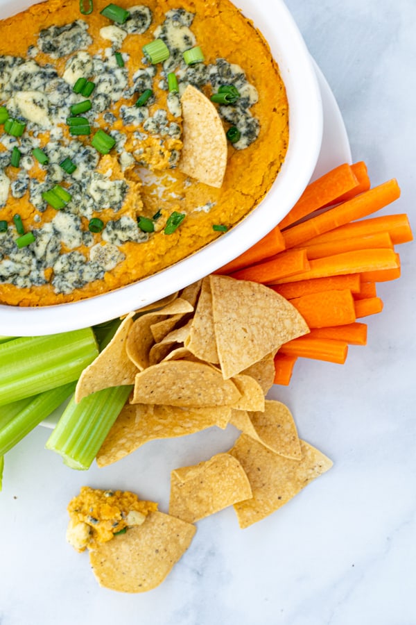 Warm Buffalo Chickpea Dip with some scooped onto a chip set down on marble background with carrots and celery
