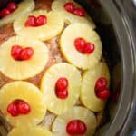 a spiral ham in the slow cooker covered by a brown sugar rub, pineapple slices, and cherries