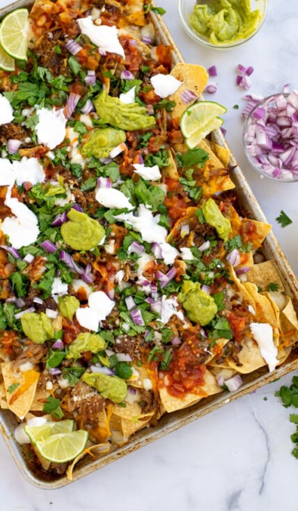 a sheet pan full of carnitas nachos topped with cilantro, red onion, guacamole, and more