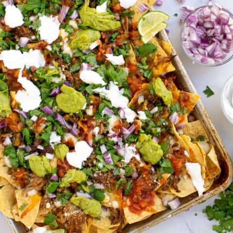 a sheet pan full of carnitas nachos topped with cilantro, red onion, guacamole, and more
