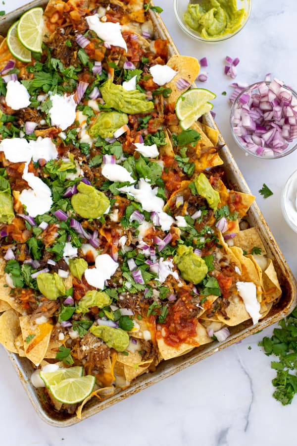 a sheet pan full of carnitas nachos topped with cilantro, red onion, guacamole, and more