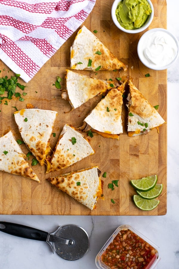 two carnitas quesadillas cut into pieces on a wooden cutting board with garnishes nearby