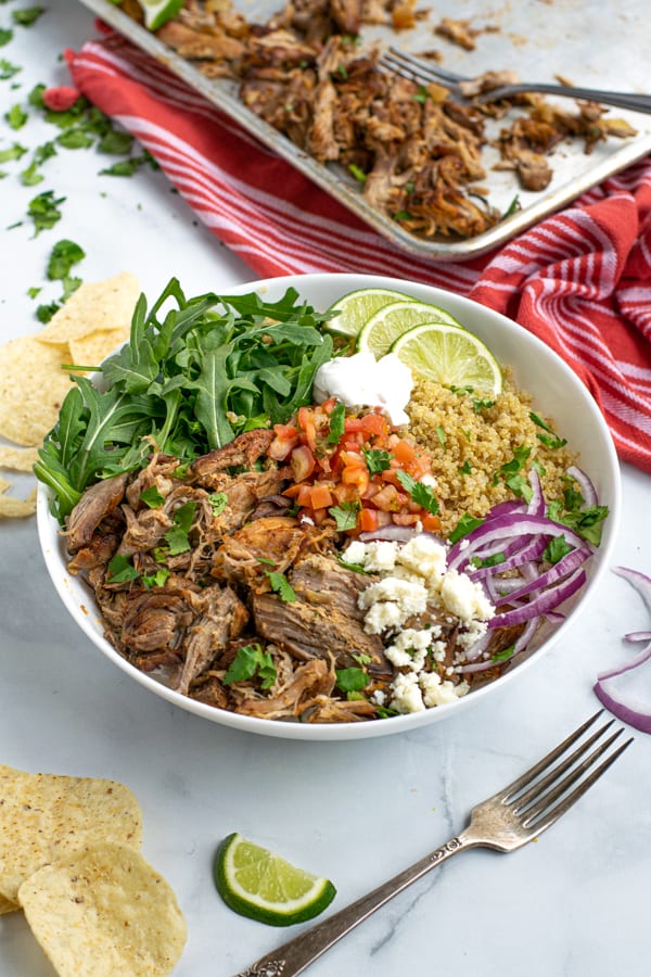 a large white bowl full of leftover carnitas, quinoa, arugula, and fresh toppings like salsa and sour cream