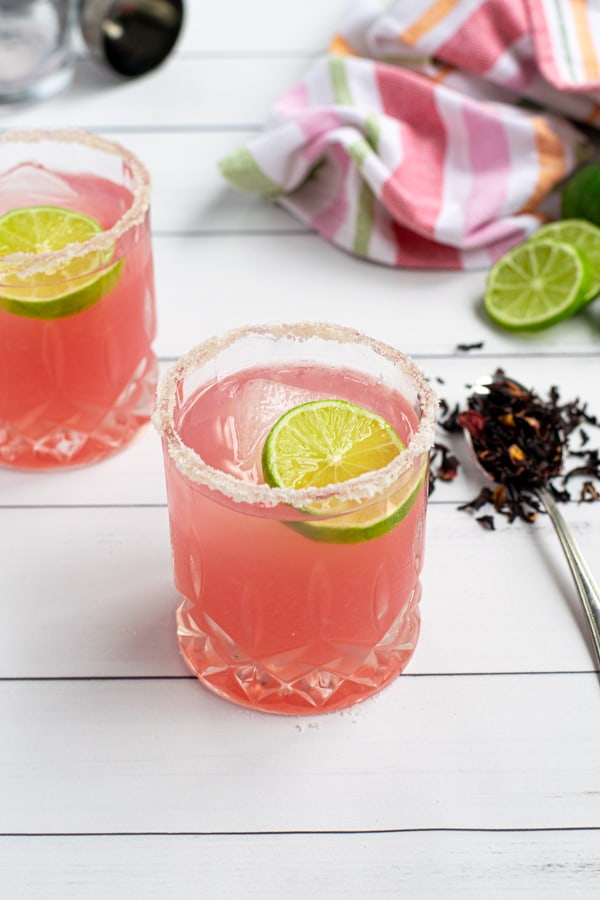 hibiscus margarita on a white table in a glass with a salted-rim and lime wheel