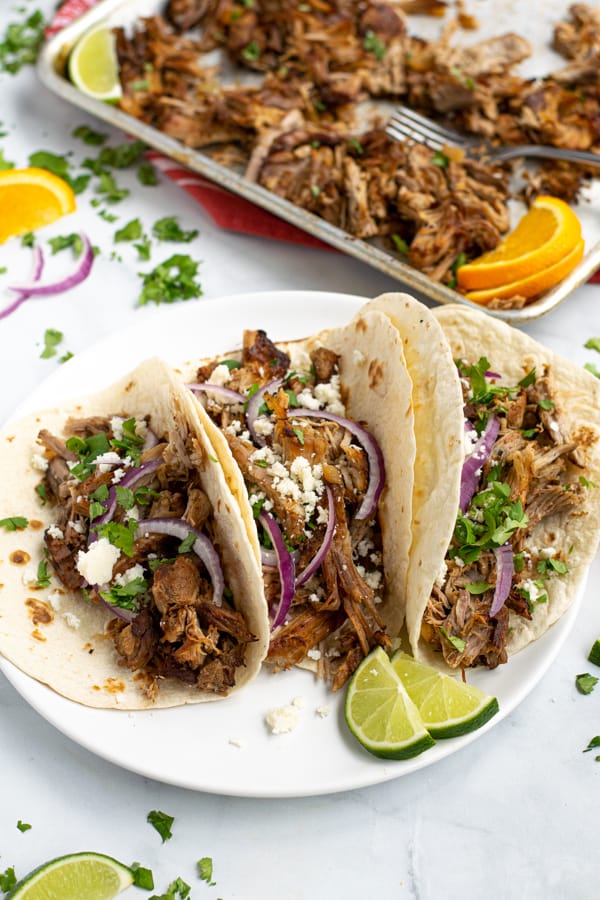 three carnitas tacos on a white plate with onions, cheese and cilantro