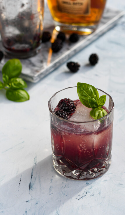 an old fashioned glass filled with a purple blackberry bourbon smash garnished with a large ice cube, blackberries, and basil